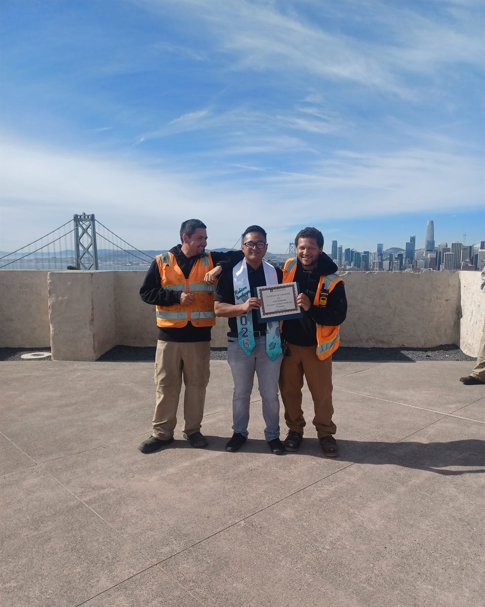 A Treasure Island landscape training graduate holding a certificate beside two Rubicon crew members, with the Bay Bridge behind them